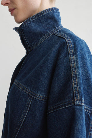 Close-up of a person wearing a dark indigo denim jacket with a high stand-up collar and contrast stitching, shown against a plain light gray background.
