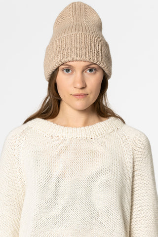 Woman in front of white background wearing ivory knit sweater and sand coloured alpaca wool handknit toque by Dinadi. 