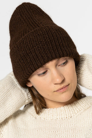 Woman wearing cream sweater and brown chunky knit toque, in front of white background. 