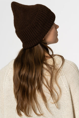 Back view of woman with long hair, wearing cream sweater and brown chunky knit toque, in front of white background. 