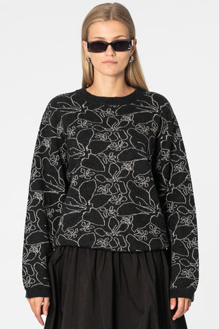 Woman in front of white wall wearing black knit crew neck sweater with grey floral design, paired with skirt and black sunglasses. 
