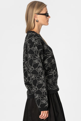Side view of woman in front of white wall wearing black knit crew neck sweater with grey floral design, paired with skirt and black sunglasses. 