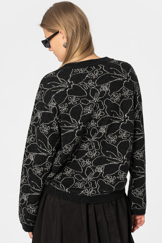Back view of woman in front of white wall wearing black knit crew neck sweater with grey floral design, paired with black skirt. 