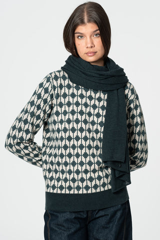 Woman standing in front of white background wearing patterned sweater and dark teal green scarf. 