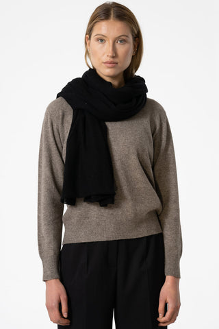 Woman standing in front of white background wearing oat brown sweater, black pants and black merino wool scarf. 
