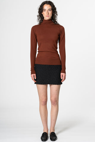 Woman in front of white background wearing brick mock neck sweater and onyx grey hand-crochet merino wool skirt by Dinadi. 