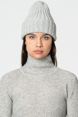 Woman in front of white background wearing grey thick rib knit toque and grey turtleneck. 