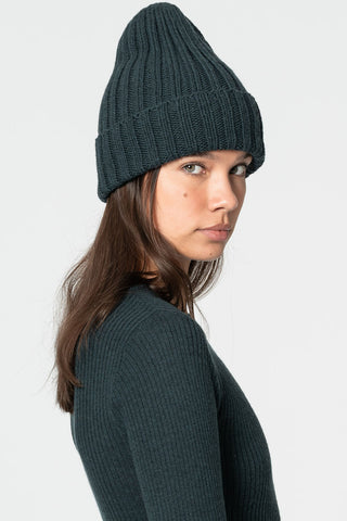 Woman in front of white background wearing dark teal thick rib knit toque and dark teal sweatshirt. 