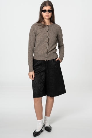 Woman in front of grey background wearing light brown yak wool knit cardigan by Dinadi paired with sunglasses, black knee length shorts, white socks and pointed toe flat shoes. 