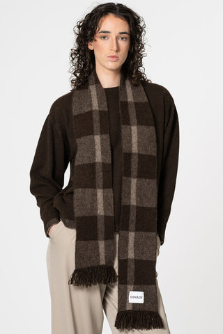 Woman standing in front of white background wearing brown sweater and brown tone plaid Dinadi scarf. 