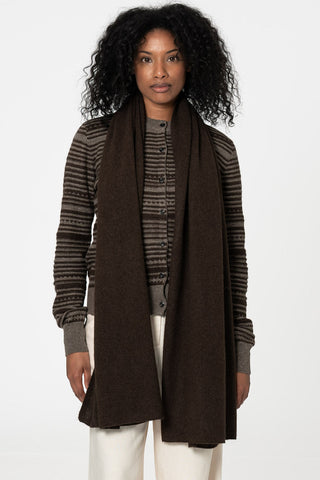 Woman standing in front of white background wearing striped cardigan, white pants and brown scarf. 