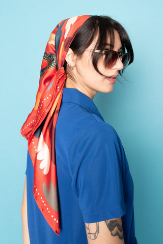 Woman in front of blue background wearing blue shirt and red & multi-colour scarf tied around her head. 