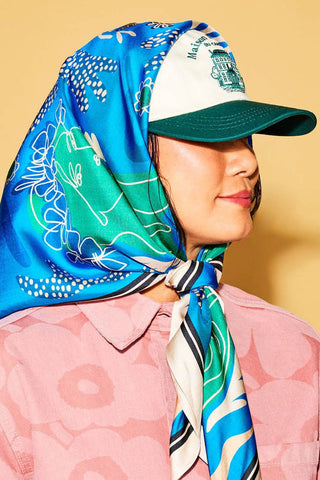Woman in front of yellow background wearing pink floral shirt, baseball cap and blue & green scarf tied around her head. 