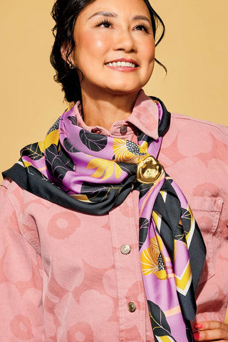 Woman in front of yellow background wearing pink floral jacket and pink, yellow and black scarf tied around her neck. 