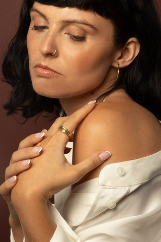 Woman in front of dark background with nose ring, gold hoop earrings and polished brass ring. 