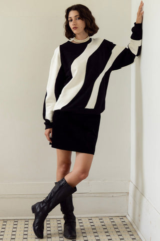 A woman wears a black and white wavy design crew neck sweater and matching black mini skirt, paired with black knee-high boots, leaning against a white wall.