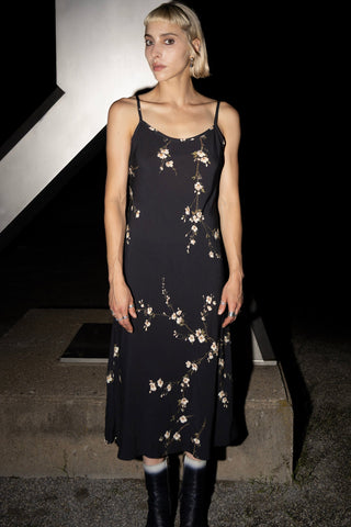 A woman wearing a black spaghetti-strap midi dress with a floral print, paired with black boots, standing against a dark background. 