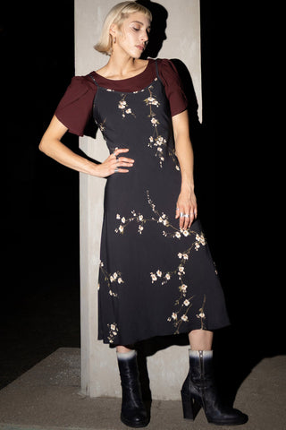 A woman wears a black midi dress with a floral pattern over a dark burgundy puff sleeves top, paired with blackboots, standing against a dark background.