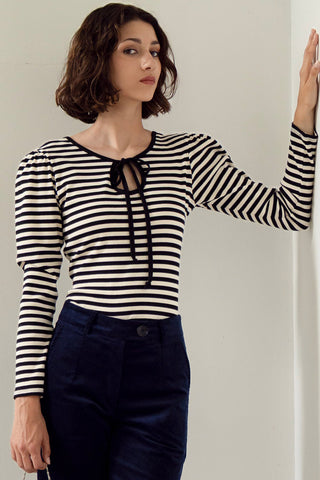 Woman standing in front of white wall wearing a black and white stripe long puff sleeve top with keyhole opening at neckline. 
