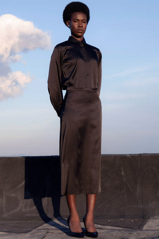 Woman stands in front of blue sky, wearing matching black satin button up shirt and a-line midi length skirt.