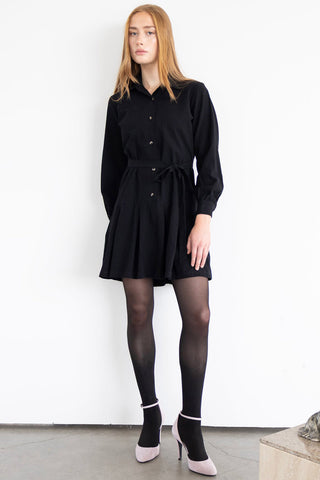 Woman in front of white wall wearing black Hope dress by Eve Lavoie, paired with black tight and mauve heels. 