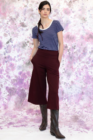 Woman in front of watercolour paint background wearing navy strip t-shirt and burgundy cropped pants. 