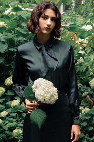 Woman standing in front of flower bushes, holding white flower, wearing dark green shirt with self-tie neck and matching skirt. 
