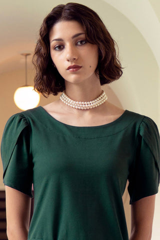 Woman standing in front of archway wearing forest green blouse with tulip style sleeves, paired with triple strand of pearls. 