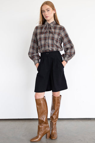 Model in front of white wall wearing plaid shirt and black knee length pleated Watson shorts by Eve Lavoie, paired with knee length brown leather boots. 