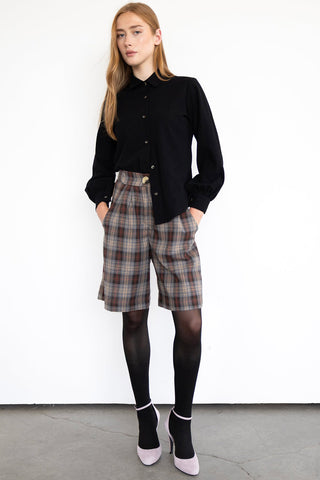 Model in front of white wall wearing black shirt and plaid knee length pleated Watson shorts by Eve Lavoie, paired with black tights and mauve heals. 