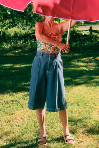 Woman in garden holding red umbrella wearing printed tank with wide leg knee length denim shorts. 