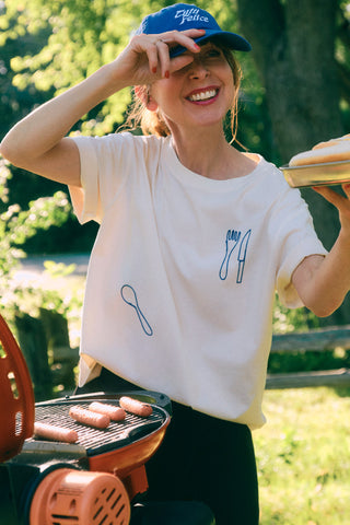 Woman grilling hot dogs wearing blue baseball cap and white t-shirt with embroidered cutlery motif. 