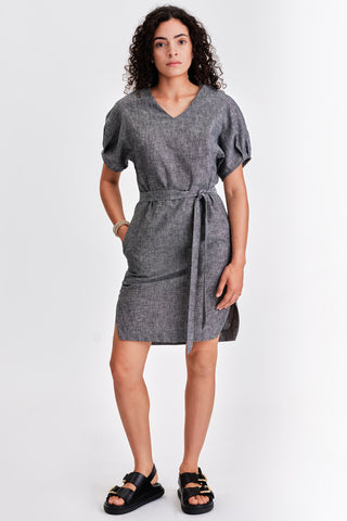 Woman in front of white background wearing dark grey belted v-neck dress with black sandals. 