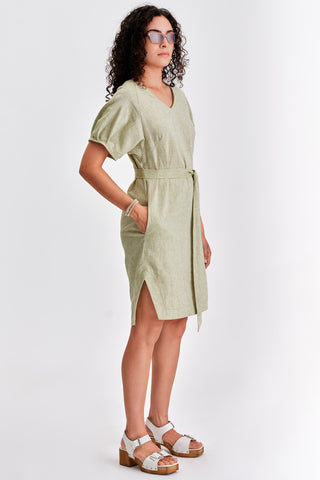 Woman in front of white background wearing light green belted v-neck dress with white sandals. 