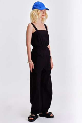 Woman in front of white background wearing blue cap and black sleeveless jumpsuit with cinched waist. 