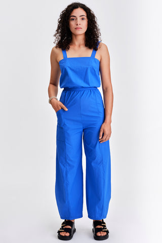 Woman in front of white background wearing bright blue sleeveless jumpsuit with cinched waist. 