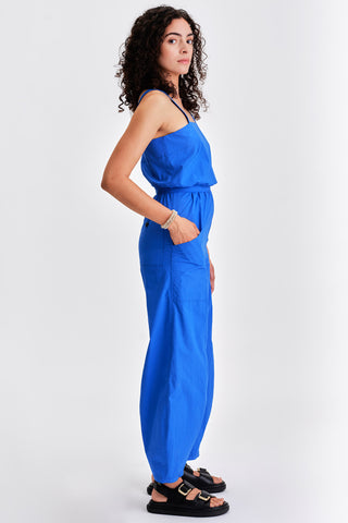 Woman in front of white background wearing bright blue sleeveless jumpsuit with cinched waist. 
