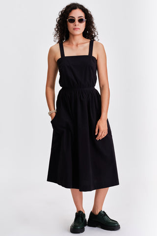 Woman in front of white background wearing sunglasses, black cinched cotton summer dress and green leather lace-up shoes. 