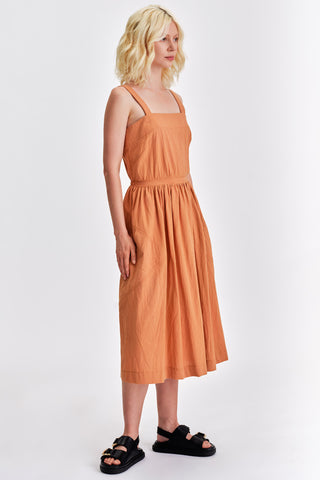 Woman in front of white background wearing  caramel orange cinched cotton summer dress and black sandals. 
