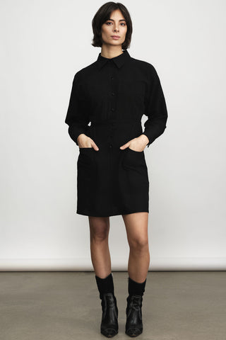A model wearing a black long-sleeve collared shirt dress with front pockets, paired with black ankle boots and socks, standing against a neutral studio background.