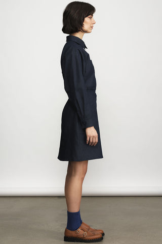 A woman in profile wearing a dark navy long-sleeve shirt dress, blue socks, and brown leather loafers, standing against a plain light gray studio background.