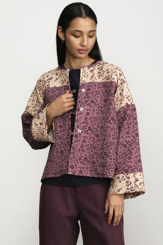 A woman wearing a cropped button-front jacket with color-blocked floral print panels in beige and dusty mauve, styled with burgundy trousers against a light gray studio background.