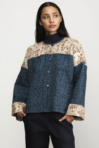 A woman wearing a cropped button-front jacket with color-blocked floral print panels in beige and blue, styled with navy trousers against a light gray studio background.