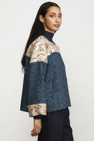 A woman wearing a cropped button-front jacket with color-blocked floral print panels in beige and blue, styled with navy trousers against a light gray studio background.