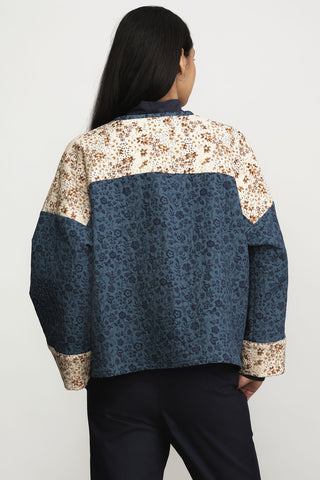 A woman shown from the back wearing a cropped jacket with color-blocked floral print panels in beige and blue, styled with navy trousers against a light gray studio background.