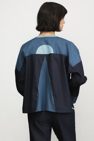A woman shown from the back wearing a denim colour-blocked jacket with "moon rising" motif, paired with dark navy pants, against a plain light gray background.