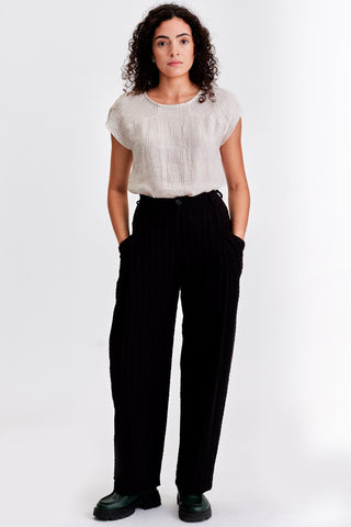 Woman in front of white background wearing ivory stripe blouse and black pleat front pants. 