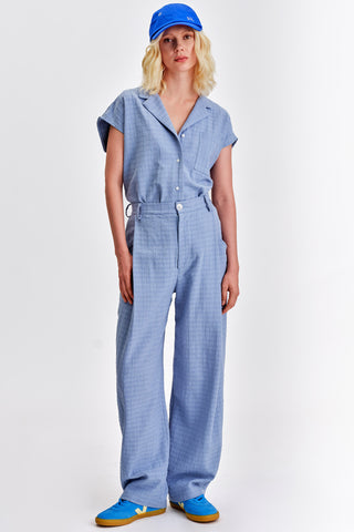 Woman in front of white background wearing blue baseball cap and matching light blue button up shirt and pleat front pants. 