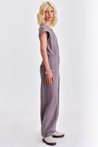 Side view of woman in front of white background wearing grey short sleeve jumpsuit with elastic waist. 