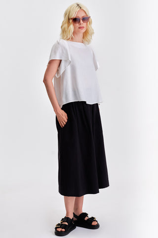 Woman in front of white background wearing white blouse, voluminous black midi length skirt and black leather sandals. 